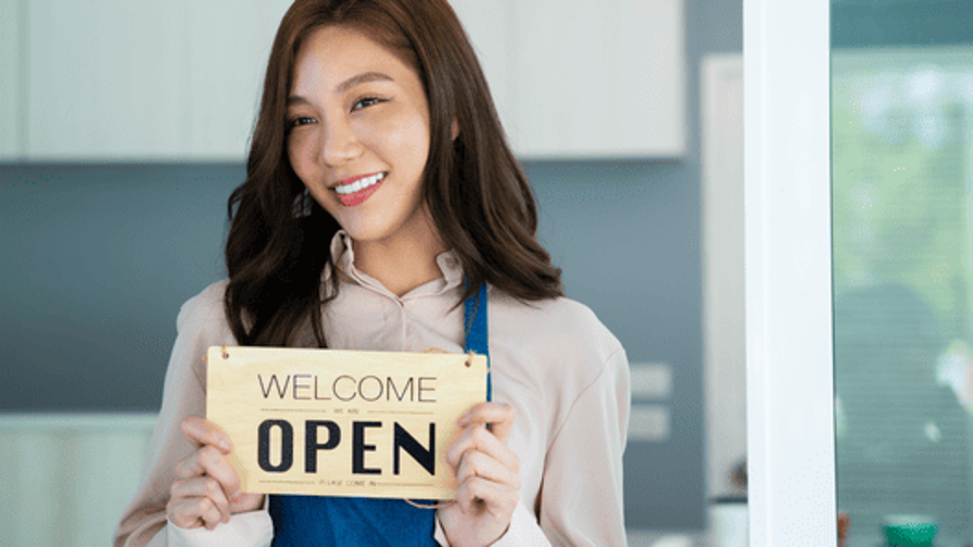 [alt=「WELCOME OPEN」と書かれたプレートをもっているカフェオーナーの姿。]