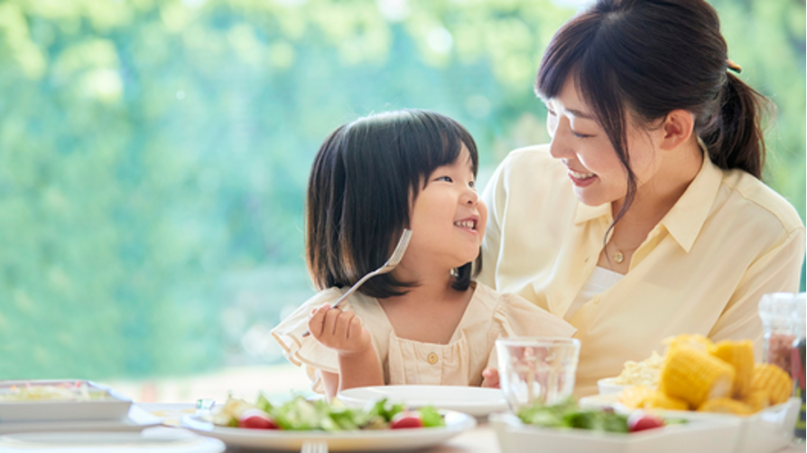 フォークを持った幼い女の子が、母親と笑顔で見つめ合いながら食事を楽しんでいる様子