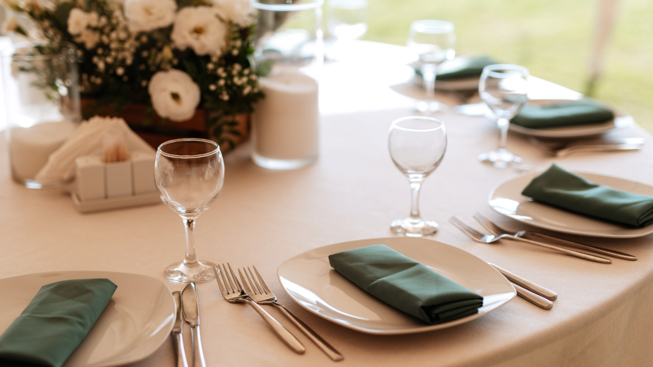 白いテーブルクロスの上に緑色のナプキンを添えたプレートとカトラリー、ワイングラスが並ぶ披露宴会場のテーブル。